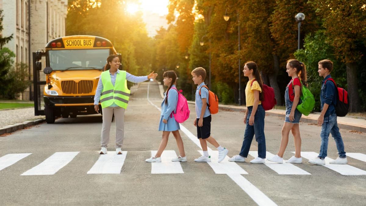 Traffic Safety: A woman in a crossing guard vest guides a group of children with colorful backpacks cross the road at a crosswalk.